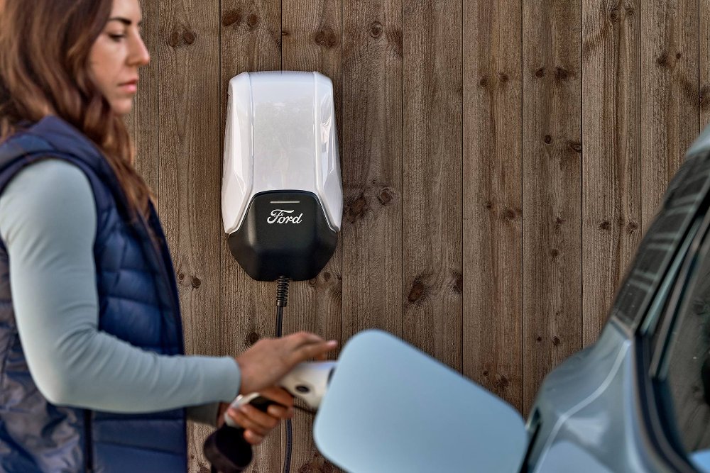 Egy nő kék Ford Explorer elektromos járművét csatlakoztatja egy otthoni elektromos járműtöltő ponthoz.