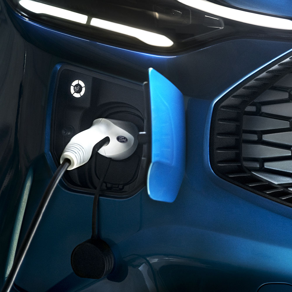 Egy kék Ford járművel szemben egy elektromos autó töltőportját látjuk, egy elektromos autótöltő csatlakozik hozzá.