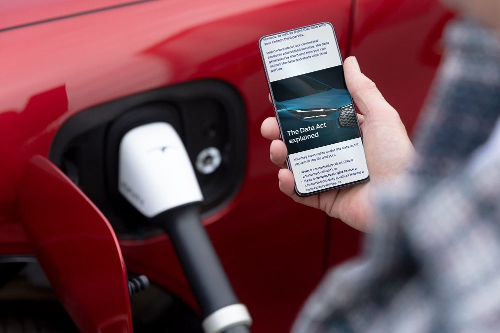 Egy személy az okostelefonjával van elfoglalva, a háttérben egy vörös színű elektromos jármű töltődik.