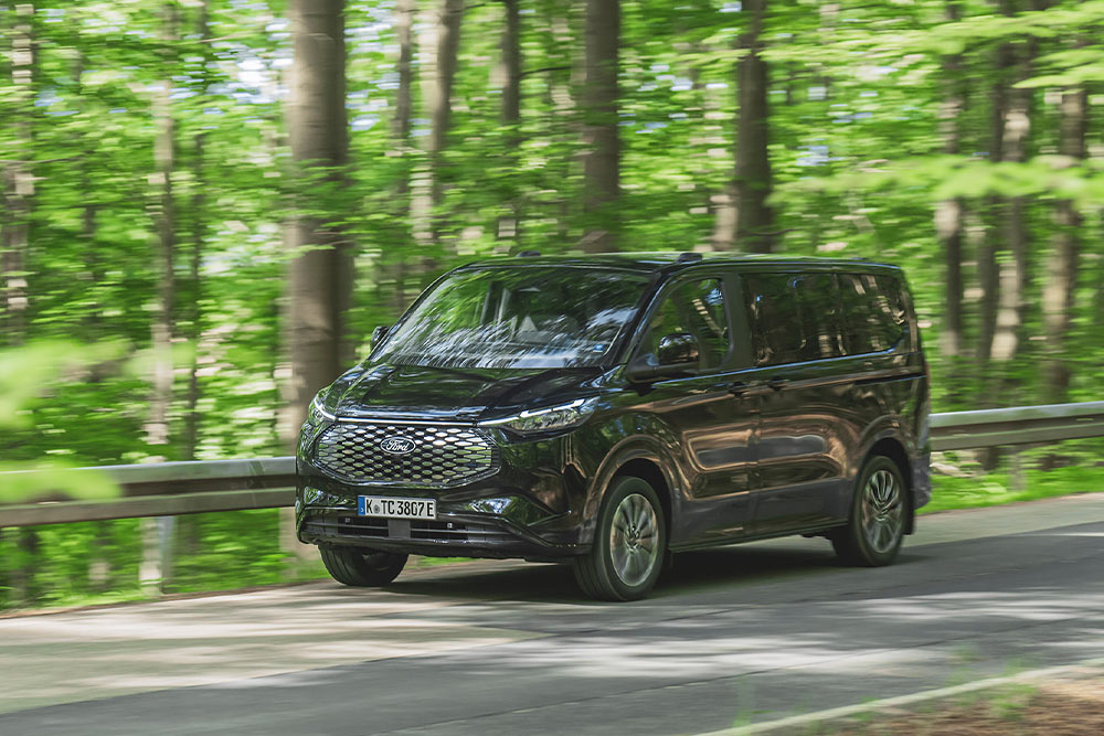 Egy fekete Ford E-Tourneo Custom egy fákkal övezett úton halad.
