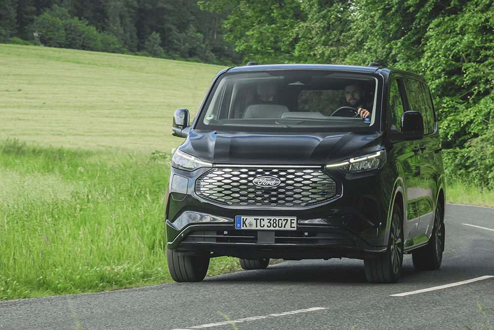 Egy fekete Ford E-Tourneo Custom vidéken halad egy úton.