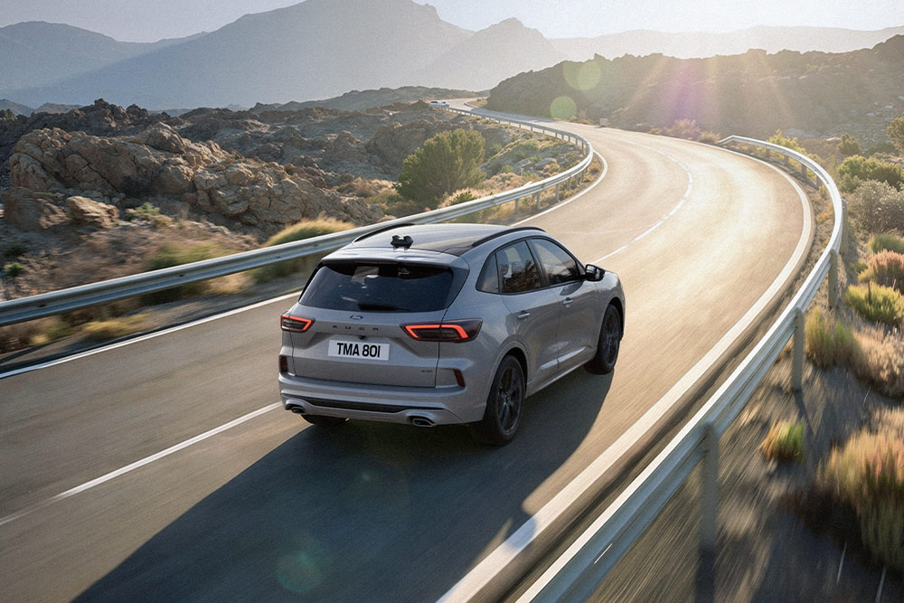 Ezüstszínű Ford Kuga egy festői úton halad, a háttérben napsütötte hegyvidéki tájjal.