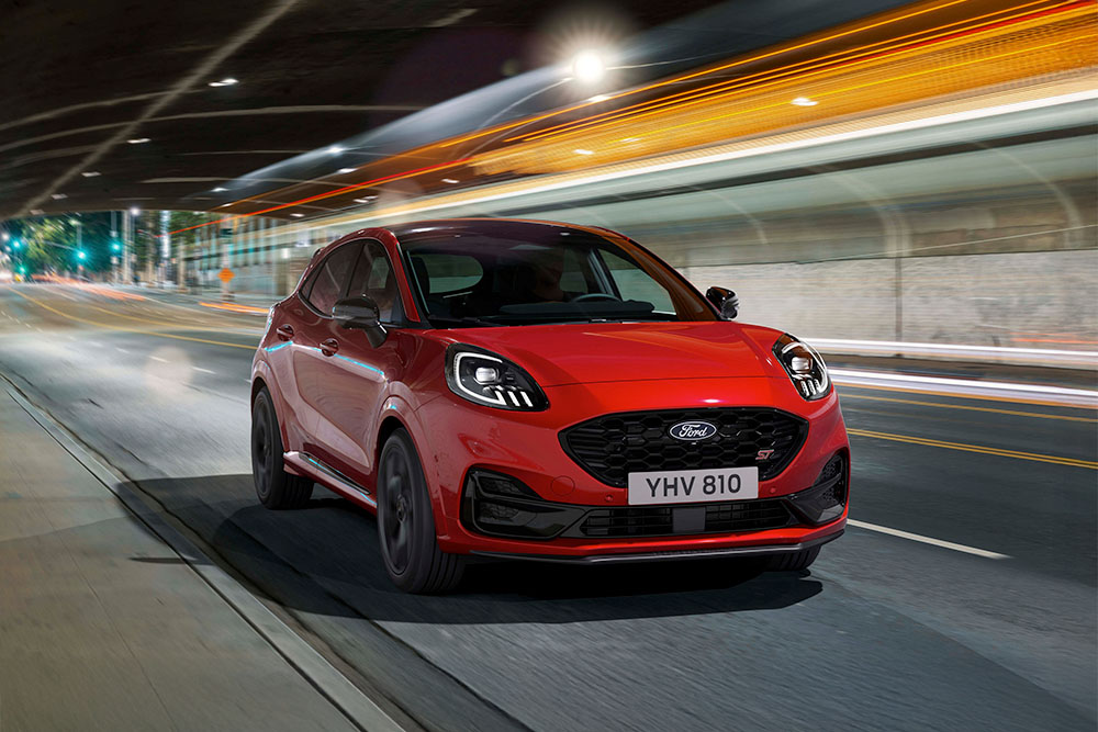 Egy piros Ford Puma ST sportos SUV, amint éjszaka egy fényesen kivilágított városi úton halad.