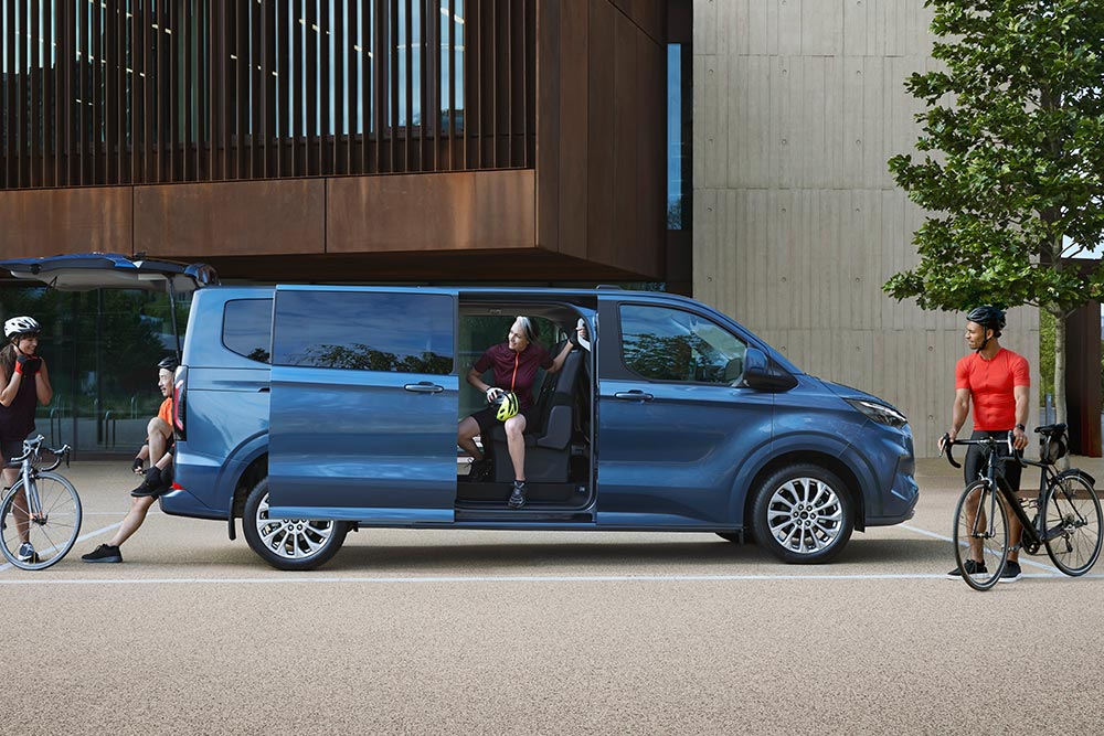 Egy kék Ford Tourneo Custom nyitott oldalsó ajtókkal, a hátuljánál kerékpárosok.