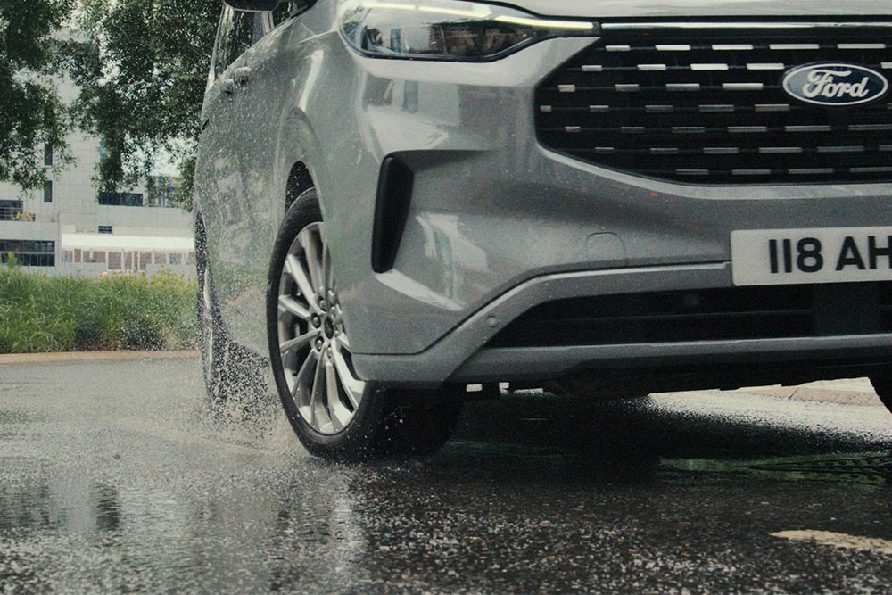 Egy ezüstszínű Ford Tourneo Custom első kereke, amint áthajt egy pocsolyán.