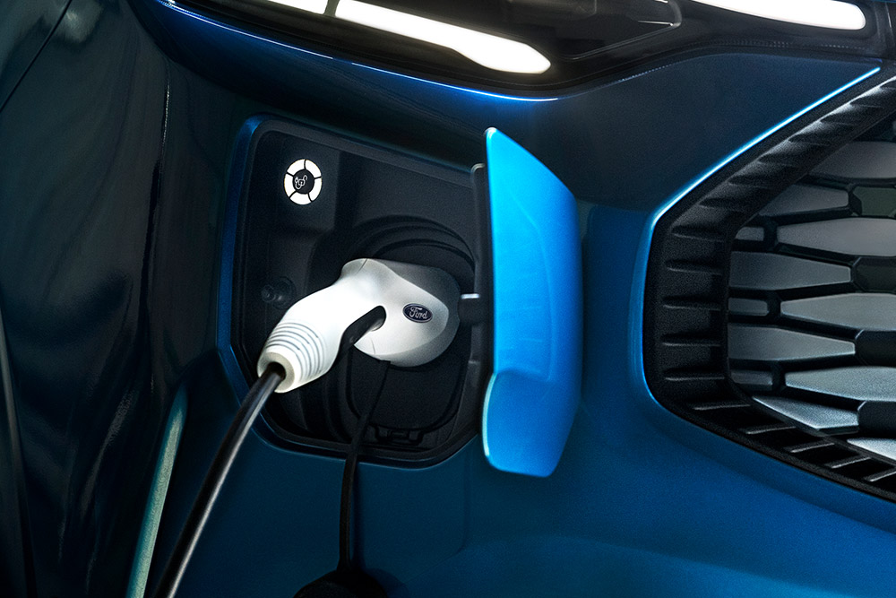Közelkép egy kék Ford E-Transit áruszállító töltőcsatlakozójáról, csatlakoztatott töltőkábellel.