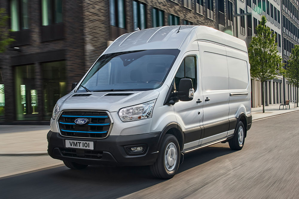 Egy ezüstszínű Ford E-Transit áruszállító egy városban halad az úton.