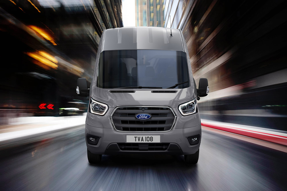 Egy szürke Ford Transit elölnézetből, amint az utcán halad. 