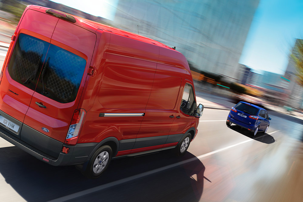 Egy piros Ford Transit Van áruszállító halad egy úton. 
