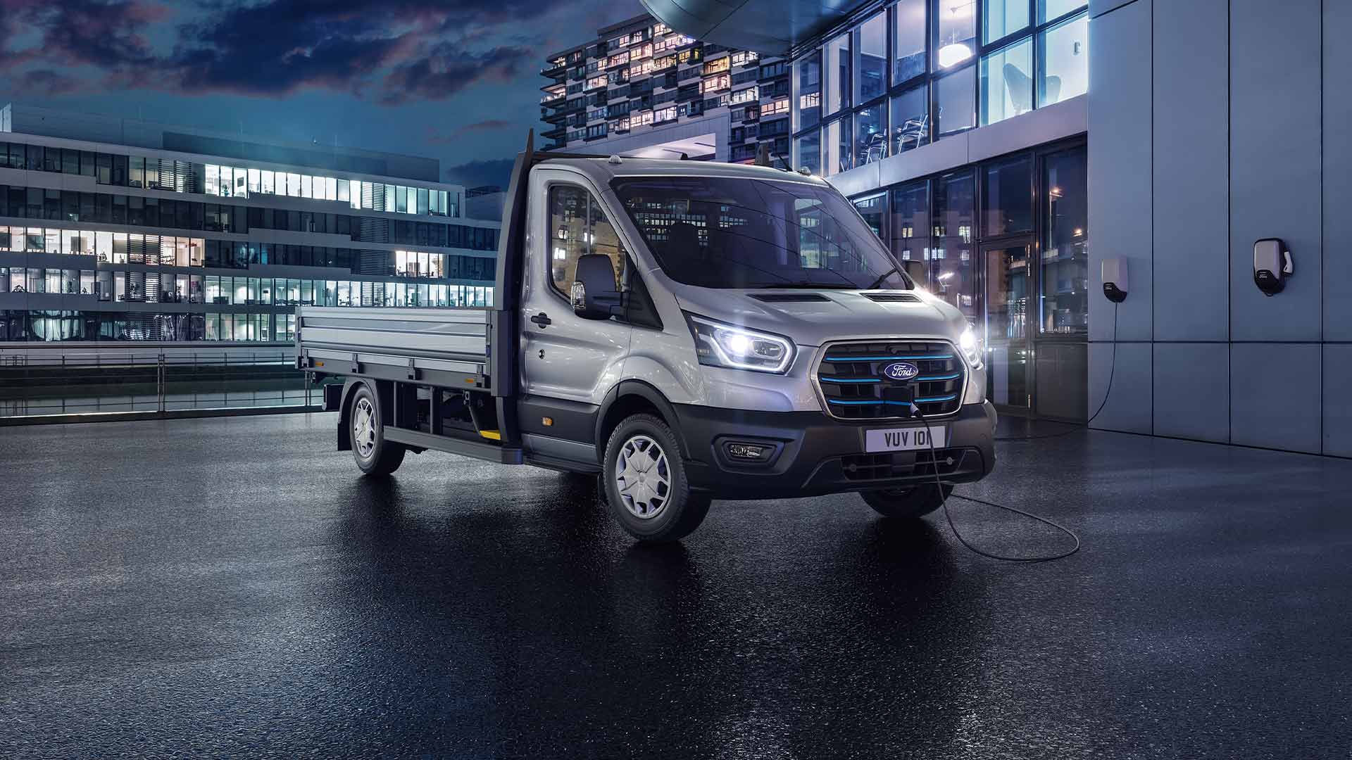Ezüstszínű Ford Transit fülkés alvázas modell parkol éjszaka egy modern épület előtt.