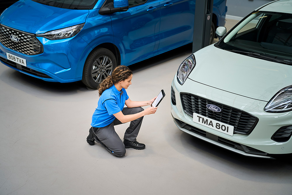 Egy technikus térdel egy kék Ford áruszállító és egy fehér Ford Puma között, és egy táblagépet néz.
