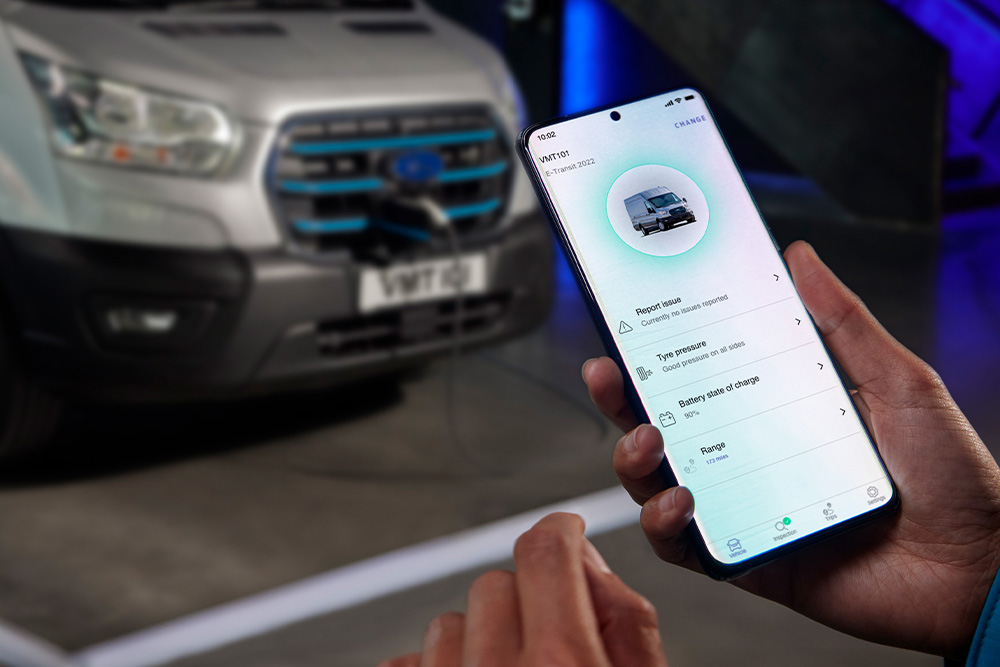 Egy személy okostelefont tart, amely Ford alkalmazást jelenít meg, a háttérben egy Ford E-Transit Custom töltődik.