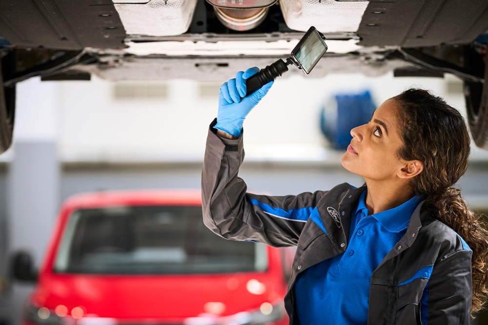 O tehniciană Ford inspectează partea inferioară a unei mașini ridicate, folosind o cameră digitală de inspecție.