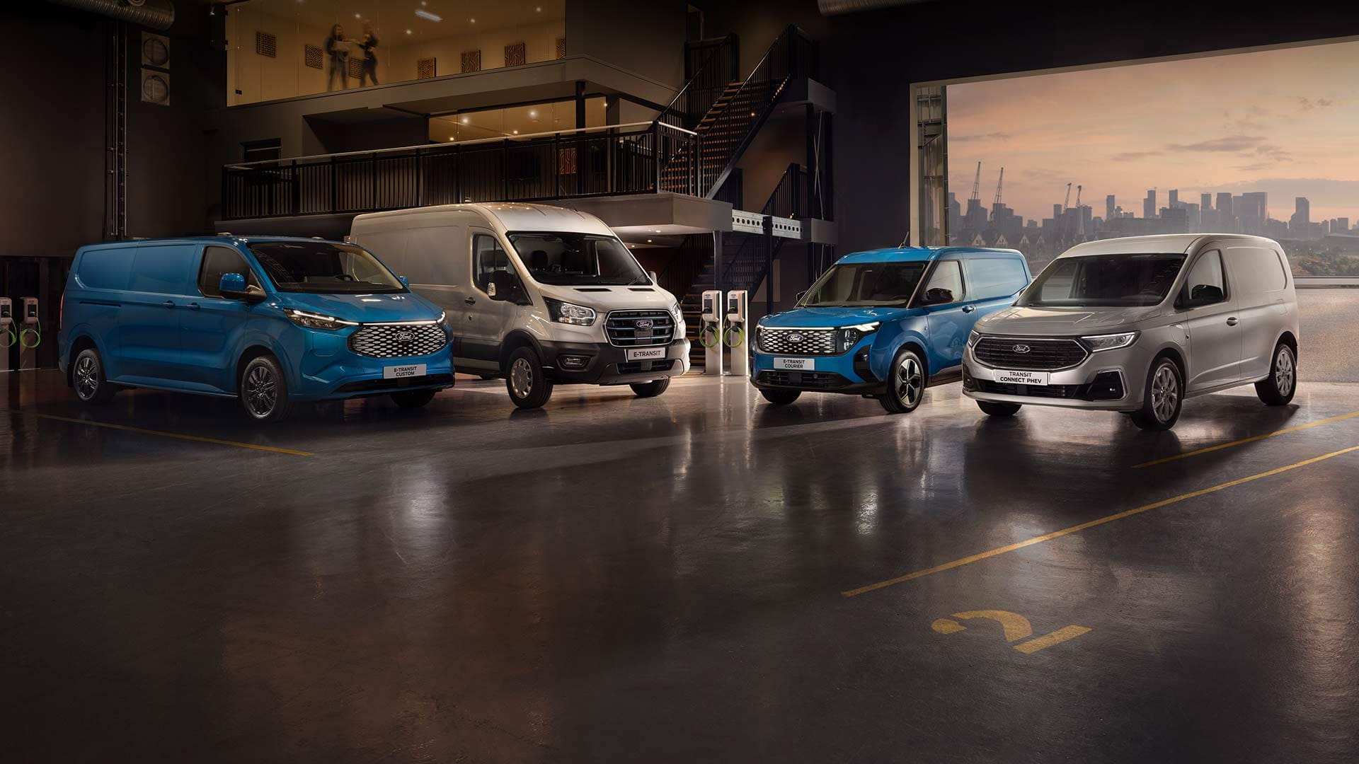 Patru furgonete electrice Ford sunt aliniate la stații de încărcare, cu un oraș vizibil în fundal.